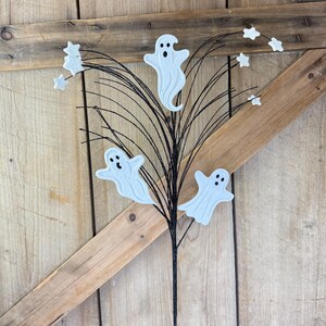 May include: A decorative floral pick featuring three white ghost cutouts with black eyes and mouths, interspersed with small white star accents on thin, flexible black wire branches that extend outward from a central black stem.