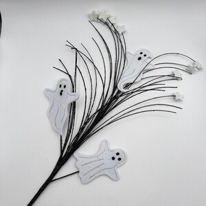 May include: A decorative floral spray featuring thin black wire branches adorned with three white ghost cutouts and several small white star-shaped accents. The ghosts have simple black circular eyes and mouths, and the entire arrangement is set against a plain white background.