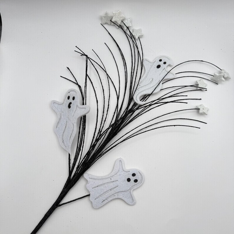 May include: A decorative floral spray featuring thin black wire branches adorned with three white ghost cutouts and several small white star-shaped accents. The ghosts have simple black circular eyes and mouths, and the entire arrangement is set against a plain white background.