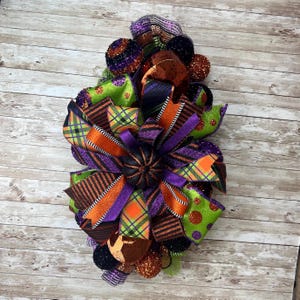 May include: A festive Halloween wreath with a variety of ribbons in orange, purple, green, and black. The wreath features plaid, polka dot, and striped patterns, along with decorative pom-poms. This handmade wreath is perfect for seasonal home decor.