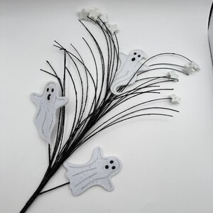 May include: A decorative floral spray featuring thin, black, wire-like stems adorned with three white, flat ghost cutouts with black facial features and several small, white, glittery star-shaped accents attached to the tips of the branches.
