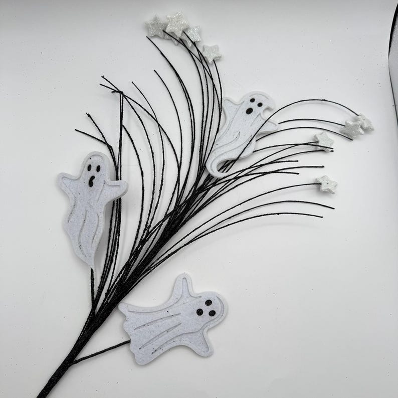May include: A decorative floral spray featuring thin, black, wire-like stems adorned with three white, flat ghost cutouts with black facial features and several small, white, glittery star-shaped accents attached to the tips of the branches.