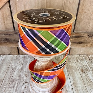 May include: A spool of decorative ribbon with a plaid pattern in orange, purple, green, black, and white. The ribbon is displayed on a white pedestal. The spool has the text "D & LUX RIBBON".