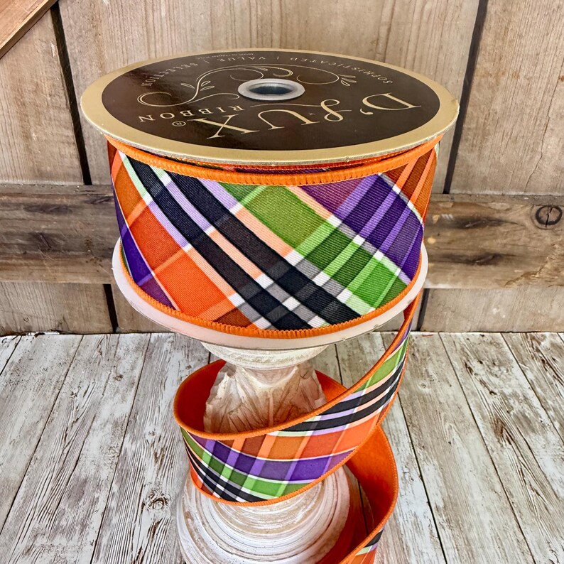 May include: A spool of decorative ribbon with a plaid pattern in orange, purple, green, black, and white. The ribbon is displayed on a white pedestal. The spool has the text "D & LUX RIBBON".