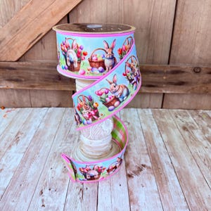 May include: A spool of light blue wired ribbon featuring a repeating pattern of brown rabbits, woven baskets filled with colourful Easter eggs, and pink and red tulips. The ribbon edges are finished with a bright pink trim and a green plaid backing.