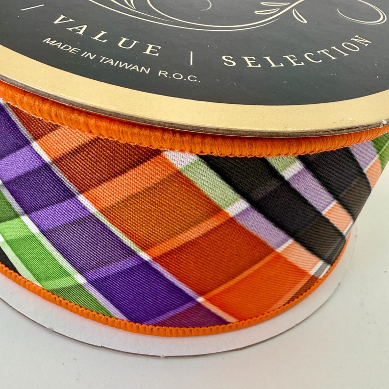 May include: A roll of decorative ribbon with a plaid pattern in orange, purple, green, and black. The ribbon is trimmed with orange edging and is labeled "VALUE | SELECTION" and "MADE IN TAIWAN R.O.C."