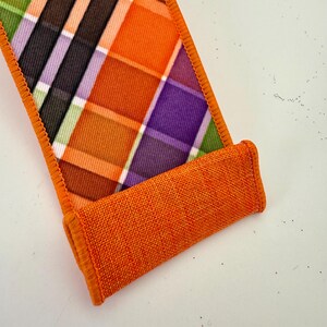 May include: A decorative ribbon with a plaid pattern in orange, purple, black, and green. The ribbon has a rolled-up section in a solid orange color, and the edges are finished with orange stitching. The ribbon is likely used for crafts or gift wrapping.