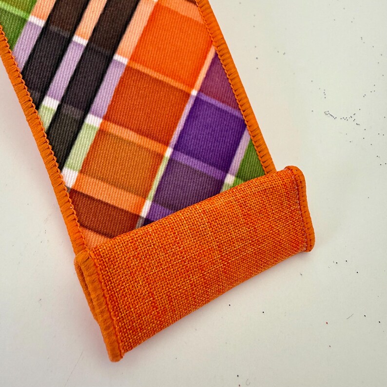 May include: A decorative ribbon with a plaid pattern in orange, purple, black, and green. The ribbon has a rolled-up section in a solid orange color, and the edges are finished with orange stitching. The ribbon is likely used for crafts or gift wrapping.