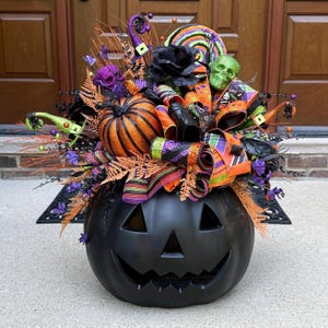 May include: A large black plastic jack-o'-lantern base topped with a decorative arrangement featuring an orange and black striped pumpkin, a green skull, a purple skull, a black rose, patterned ribbons, and miniature witch hats.