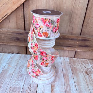 May include: A spool of wired ribbon featuring a vibrant floral pattern with pink, orange, and red roses on a light beige background. The ribbon is wrapped around a cylindrical spool and cascades downward in a spiral, showcasing the detailed botanical print.