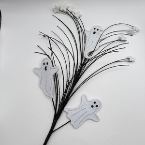 May include: A decorative floral spray featuring thin, flexible black stems adorned with three white, glittery ghost cutouts and several small, white glittery star accents attached to the tips of the stems, creating a festive Halloween arrangement.