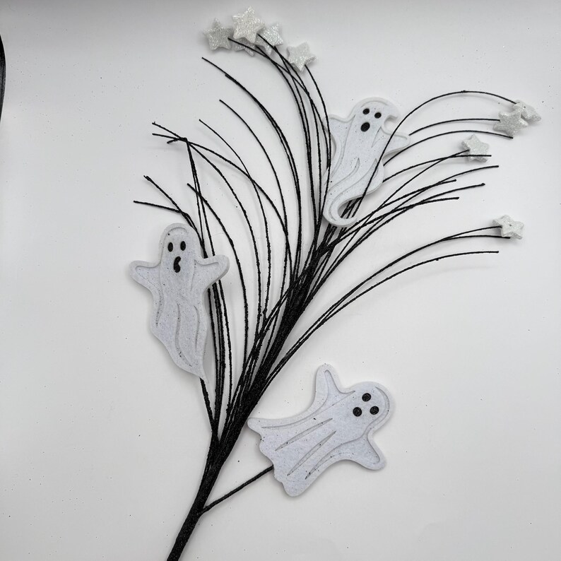 May include: A decorative floral spray featuring thin, flexible black stems adorned with three white, glittery ghost cutouts and several small, white glittery star accents attached to the tips of the stems, creating a festive Halloween arrangement.