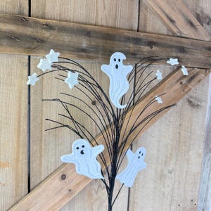 May include: A decorative floral spray featuring three white ghost cutouts with black eyes and mouths, interspersed with small white glittery stars attached to thin, flexible black wire stems that fan outward from a central base point.