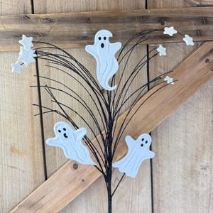 May include: A decorative floral pick featuring three white ghost cutouts with black eyes and mouths, interspersed with small white glittery star shapes attached to thin, flexible black wire stems that fan outward from a central base.