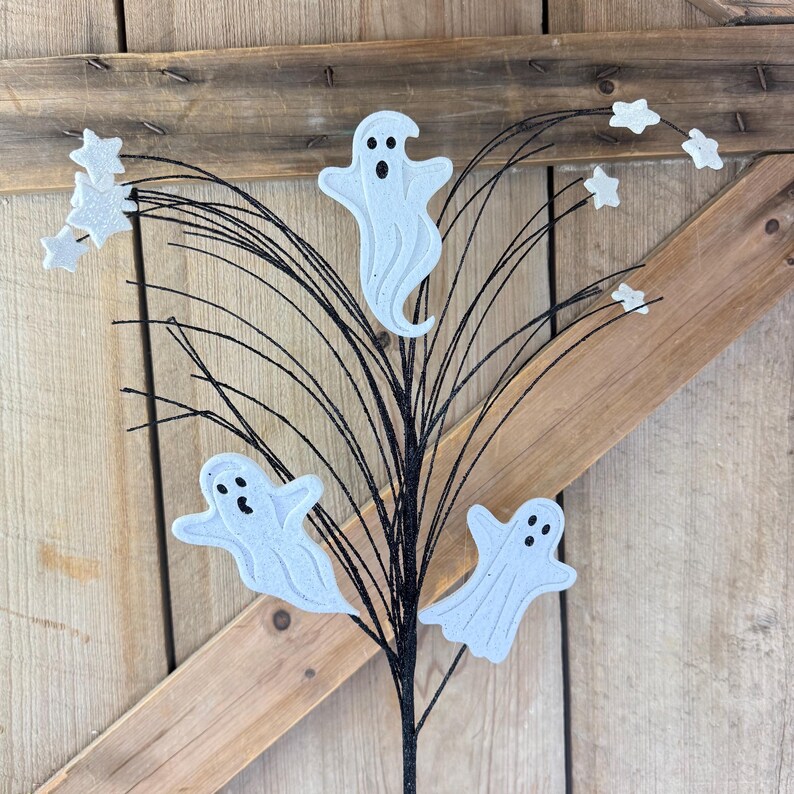 May include: A decorative floral pick featuring three white ghost cutouts with black eyes and mouths, interspersed with small white glittery star shapes attached to thin, flexible black wire stems that fan outward from a central base.
