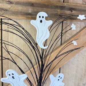 May include: A decorative spray featuring three white ghost cutouts with black eyes and mouths, attached to thin, flexible black wire stems. Several small white star-shaped accents are interspersed among the black wire branches.