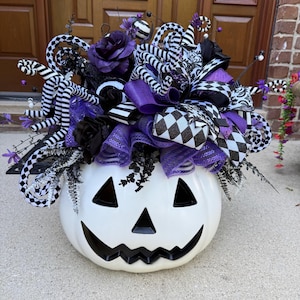 May include: A white jack-o'-lantern pumpkin decoration topped with a large arrangement of purple roses, black roses, black and white striped ribbons, and checkered patterned ribbons. The display features curly wire accents and purple mesh.