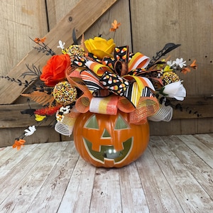 May include: A decorative orange jack-o'-lantern pumpkin centerpiece featuring a vibrant arrangement of orange and yellow fabric flowers, black leafy stems, and a multi-layered bow with candy corn patterns and glittery orange and white mesh ribbons.