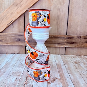 May include: A roll of white fabric ribbon featuring a Halloween-themed pattern of orange and grey pumpkins, black crows, and spider webs. The ribbon has a bright orange stitched edge and is wrapped around a white, tiered, pedestal-style holder.