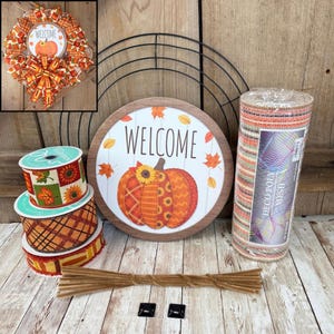May include: A DIY wreath making kit featuring a black wire frame, a round wooden sign with a pumpkin and the text WELCOME, three rolls of patterned ribbon with autumn leaves and plaid designs, a roll of deco poly mesh, and brown pipe cleaners.