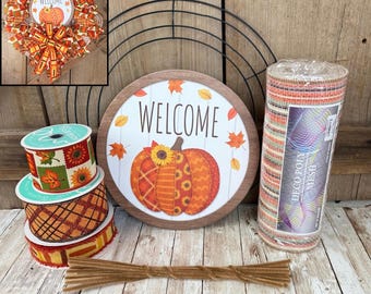 May include: A DIY wreath making kit featuring a black wire frame, a round wooden sign with a pumpkin and the text WELCOME, three rolls of patterned ribbon with autumn leaves and plaid designs, a roll of deco poly mesh, and brown pipe cleaners.