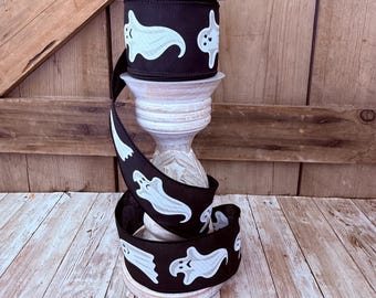 May include: A roll of black fabric ribbon featuring a repeating pattern of white cartoon ghosts with simple facial expressions. The ribbon is wrapped around a white decorative pedestal, displaying the ghost print along its entire length.
