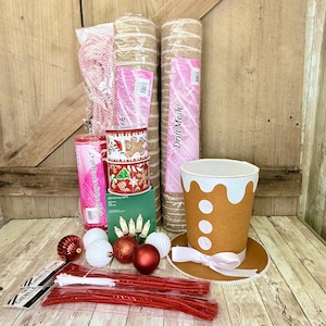 May include: A craft supply set featuring two rolls of tan mesh, a roll of pink mesh, a roll of red and white striped cord, two spools of gingerbread-themed ribbon, a box of string lights, red and white ornaments, and a gingerbread hat.