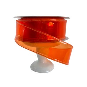 May include: A spool of bright orange sheer ribbon with a wired edge. The ribbon is displayed on a white stand.
