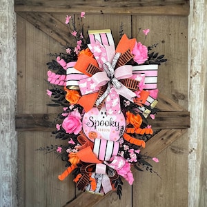 May include: A Halloween-themed wreath with pink, orange, and black accents. The wreath features a pink pumpkin-shaped sign that reads "Spooky Season" and is decorated with ribbons, flowers, and ghost designs. The wreath is displayed on a wooden door.
