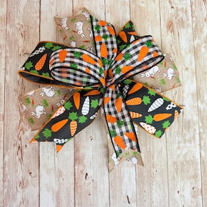 May include: An Easter-themed bow with various ribbon patterns. The bow incorporates black ribbons with orange carrot designs, black and white gingham ribbons, and tan ribbons with white bunny prints. A colourful decoration for the Easter season.