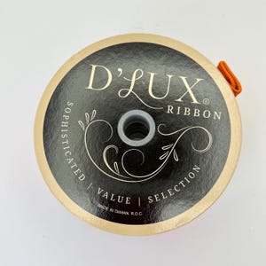 May include: A spool of D'Lux Ribbon with a black and gold label. The label features the text "D'Lux Ribbon" and decorative flourishes. The spool has an orange ribbon loop attached. The words "Sophisticated", "Value", and "Selection" are also printed on the label.