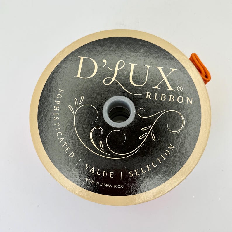 May include: A spool of D'Lux Ribbon with a black and gold label. The label features the text "D'Lux Ribbon" and decorative flourishes. The spool has an orange ribbon loop attached. The words "Sophisticated", "Value", and "Selection" are also printed on the label.