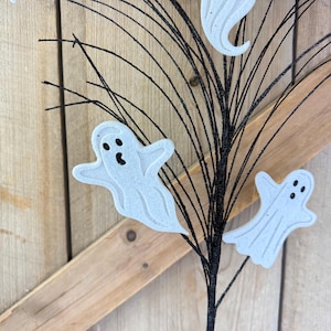May include: A decorative spray featuring thin, black, glittery branches adorned with three white, flat ghost cutouts. Each ghost has a simple, embossed design with two black eyes and a small black mouth, attached to the dark, twig-like stems.