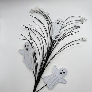 May include: A decorative spray featuring three white felt ghost cutouts with black eyes and mouths, attached to thin black wire stems. The arrangement is accented with small, white glittery star shapes at the ends of several wire branches.