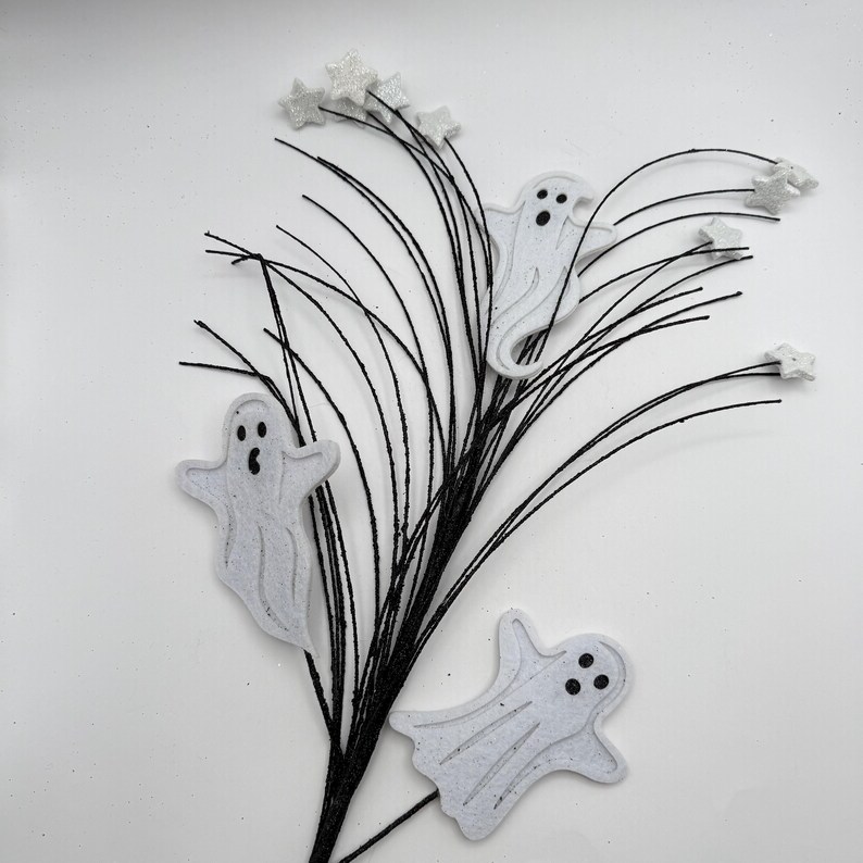 May include: A decorative spray featuring three white felt ghost cutouts with black eyes and mouths, attached to thin black wire stems. The arrangement is accented with small, white glittery star shapes at the ends of several wire branches.