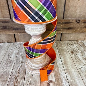 May include: A decorative ribbon with a plaid pattern in orange, purple, green, black, and white. The ribbon is displayed on a white, wooden pedestal. The ribbon's edges are orange, and it is likely used for crafting or decorating.