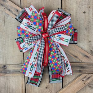 May include: A large decorative bow with multiple ribbon patterns. The bow features red, green, and blue plaid, black and white checkered, and ribbons with nutcracker soldier designs. The bow is tied with a red center and is set against a wooden background.