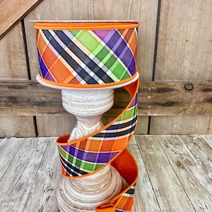 May include: A roll of decorative ribbon with a plaid pattern in orange, purple, green, black, and white. The ribbon is displayed on a white, wooden pedestal against a rustic wooden background. The ribbon's edges are trimmed with orange.