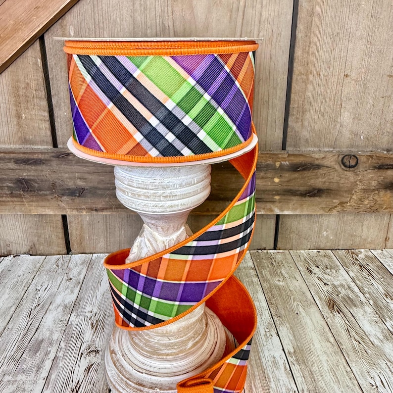May include: A roll of decorative ribbon with a plaid pattern in orange, purple, green, black, and white. The ribbon is displayed on a white, wooden pedestal against a rustic wooden background. The ribbon's edges are trimmed with orange.