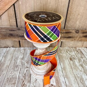 May include: A spool of decorative ribbon with a plaid pattern in orange, purple, green, and black. The ribbon is displayed on a white pedestal. The spool has the text "D'Lux Ribbon" on the top.