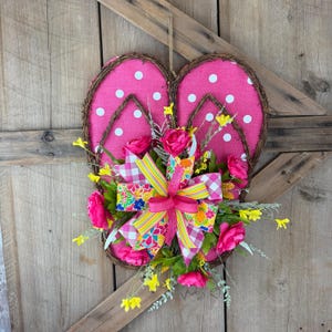 May include: A decorative flip-flop wreath with a pink polka-dot design. The wreath is adorned with pink roses, yellow flowers, and a colorful bow. The wreath is made of woven branches and is hanging on a wooden door.