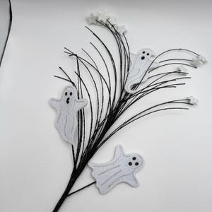 May include: A decorative spray featuring three white felt ghost cutouts with black eyes and mouths, attached to thin black wire stems. The arrangement is accented with small, white glittery star shapes at the tips of the black wire branches.