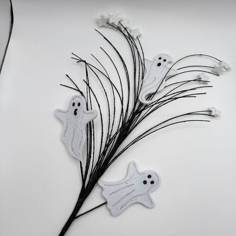 May include: A decorative spray featuring three white felt ghost cutouts with black eyes and mouths, attached to thin black wire stems. The arrangement is accented with small, white glittery star shapes at the tips of the black wire branches.