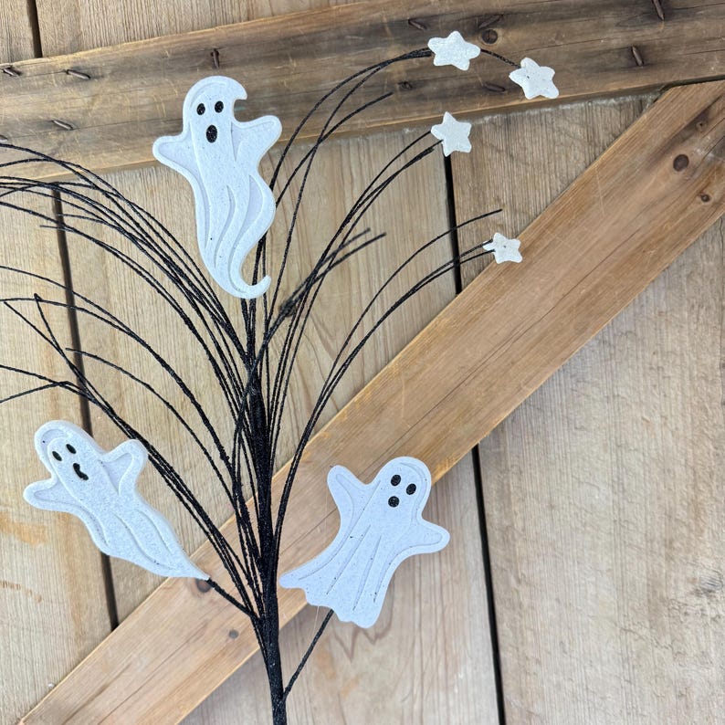 May include: A decorative floral spray featuring three white ghost cutouts with black eyes and mouths, interspersed with small white star shapes attached to thin, flexible black wire stems that fan out from a central base.