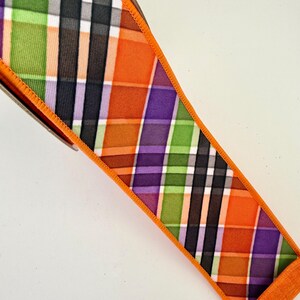 May include: A decorative ribbon with a plaid pattern in orange, green, purple, black, and white. The ribbon has an orange border and is angled diagonally across the frame. The ribbon is likely for crafting or gift wrapping.