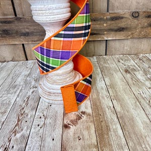 May include: A decorative orange ribbon with a plaid pattern in orange, purple, green, and black. The ribbon is wrapped around a white, wooden, decorative pillar. The background is a wooden surface.