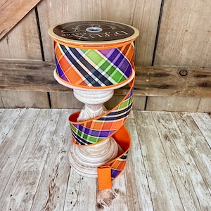 May include: A spool of decorative ribbon with a plaid pattern in orange, purple, green, black, and white. The ribbon is displayed on a white, ornate stand. The spool has the text "D.Lux Ribbon".