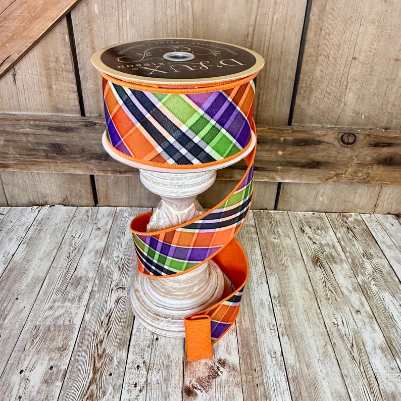 May include: A spool of decorative ribbon with a plaid pattern in orange, purple, green, black, and white. The ribbon is displayed on a white, ornate stand. The spool has the text "D.Lux Ribbon".