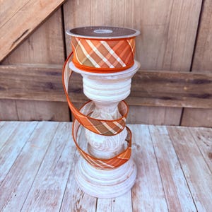 May include: A spool of orange, brown, and white plaid ribbon resting on a white distressed wooden candle holder. The ribbon features a diagonal checkered pattern and is wrapped around the base of the candle holder in a spiral design.
