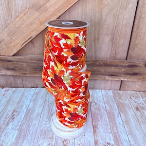 May include: A spool of wired ribbon featuring a vibrant autumn leaf pattern with shades of red, orange, yellow, and green against a white background. The ribbon has a finished orange edge and is wrapped around a circular plastic core.